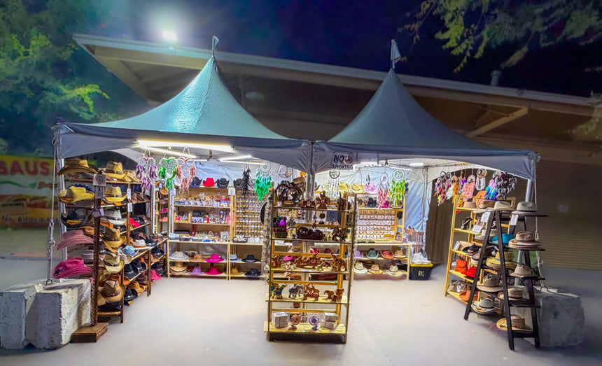 Peritik booth lit up at night at the Alameda County Fair 2026, hat shelves and natural stone bracelet wall under white canopies
