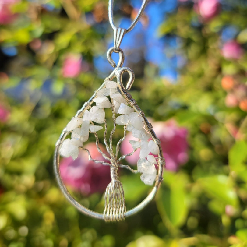 Piedra Luna, teardrop pendant with wire tree of white stone chips held outdoors against blurred pink blooms