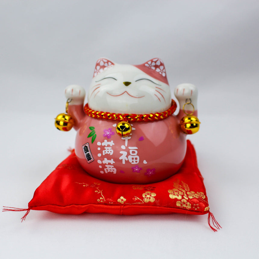 Ceramic Lucky Cat Coin Bank on Cushion, pink front view, raised paws, gold bells, painted characters, red cushion