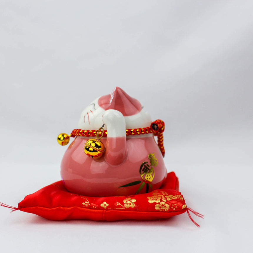 Ceramic Lucky Cat Coin Bank on Cushion, pink profile with a paw curled to its cheek, bells hanging, on the red cushion