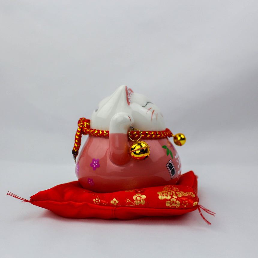 Ceramic Lucky Cat Coin Bank on Cushion, pink side view showing face markings, twin gold bells, and the tasseled red cushion
