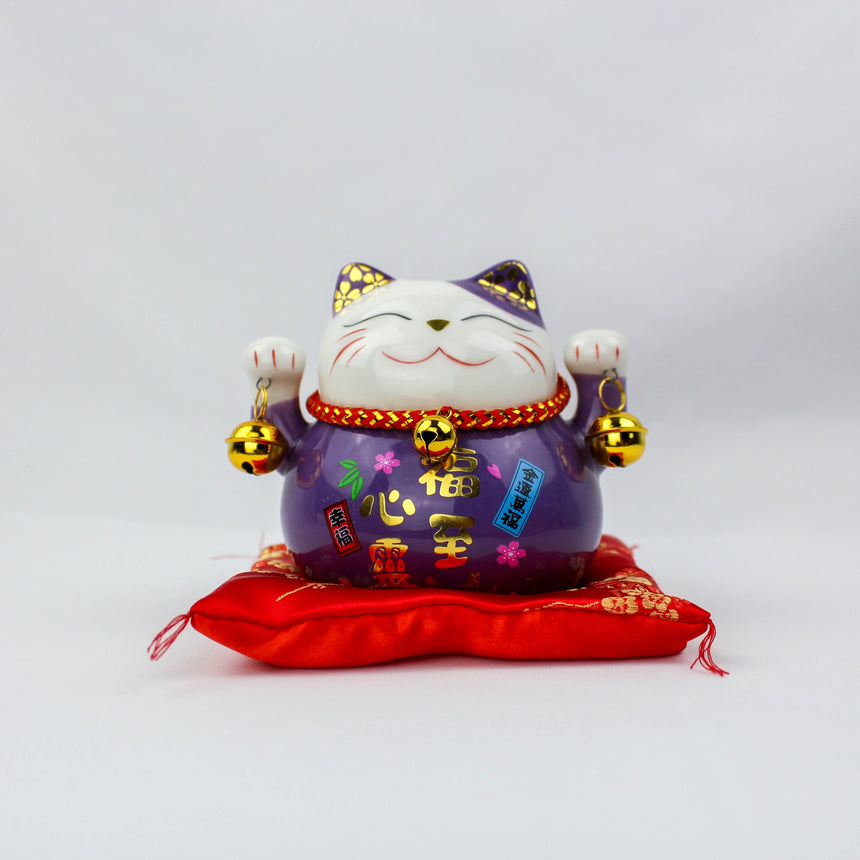Ceramic Lucky Cat Coin Bank on Cushion, purple front view with raised paws, bells, and colorful labels on a red cushion