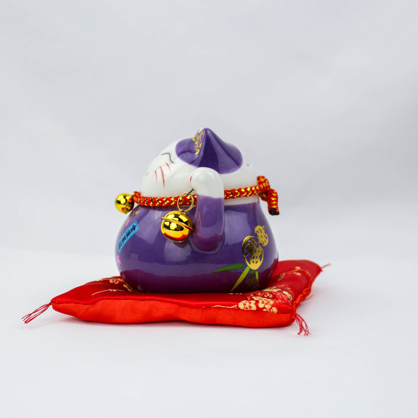 Ceramic Lucky Cat Coin Bank on Cushion, purple side profile with paw to its cheek, gold bells, and the red rope collar