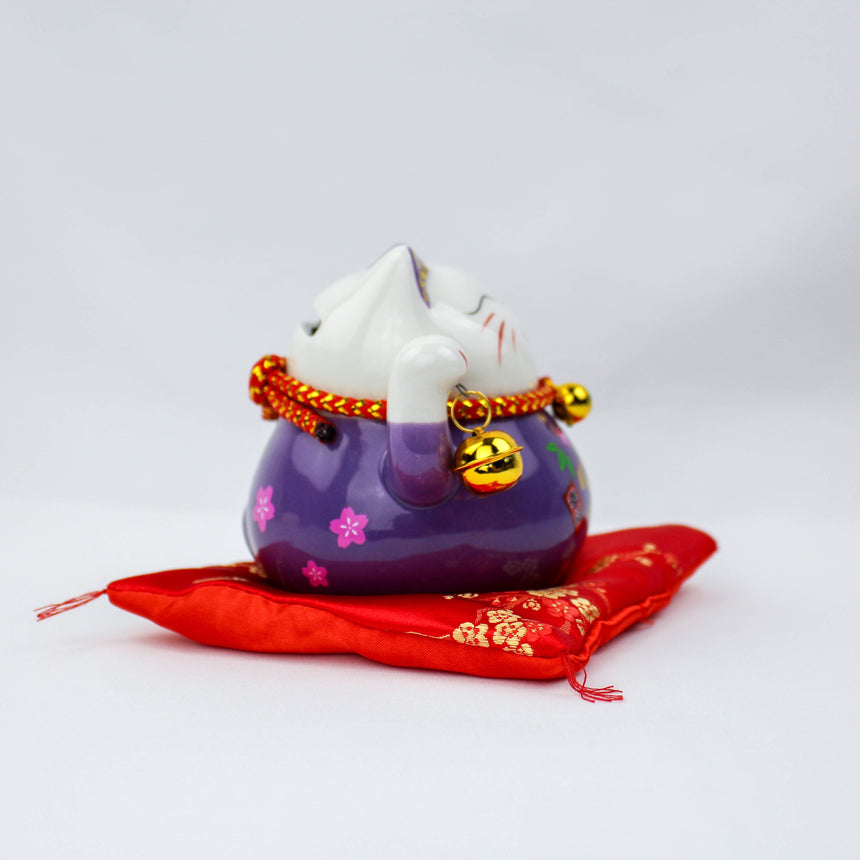 Ceramic Lucky Cat Coin Bank on Cushion, purple profile showing blossom motifs and a gold bell, resting on the red cushion